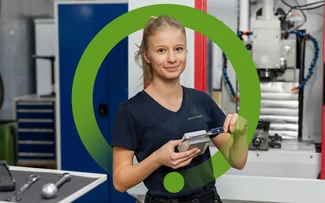 Eine Auszubildende zur Zerspanungsmechanikerin misst ein Bauteil mit einem Messschieber vor einer CNC-Fräsmaschine