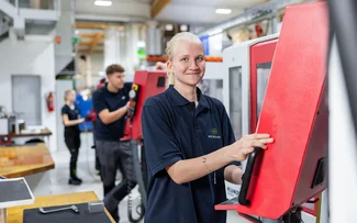 Eine Auszubildende zur Zerspanungsmechanikerin steht grinsend an der CNC-Fräsmaschine